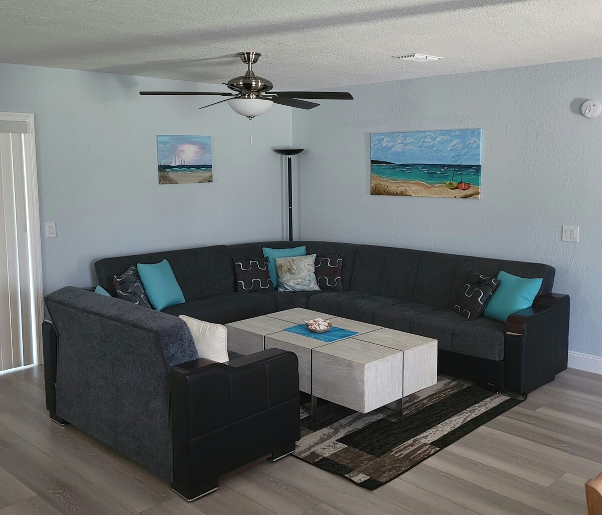 A spacious living area is furnished with a large, comfortable sectional couch adorned with decorative pillows. A coffee table sits in the center, surrounded by light-colored rugs. Coastal-themed artwork decorates the walls, adding a serene touch. A ceiling fan and natural light from nearby windows enhance the space.