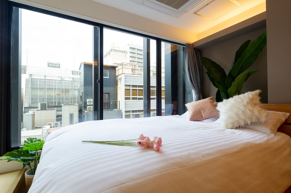 A spacious bed is positioned near large floor-to-ceiling windows, providing a view of the cityscape. A decorative arrangement of flowers rests gently on the bedding, while a green plant adds a touch of nature to the modern design. Soft lighting enhances the serene ambiance.