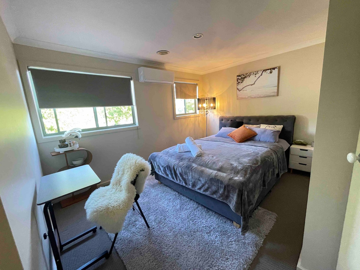 A serene guest bedroom is presented with a spacious bed dressed in soft linens and an accent pillow. A desk and chair are situated in one corner, while natural light enters through dual windows with shades.
