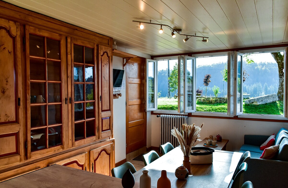 A spacious dining area features a wooden table surrounded by chairs. Large windows offer views of the outdoors, allowing natural light to enter the room. A wooden cupboard houses decorative items, enhancing the rustic charm of the space.