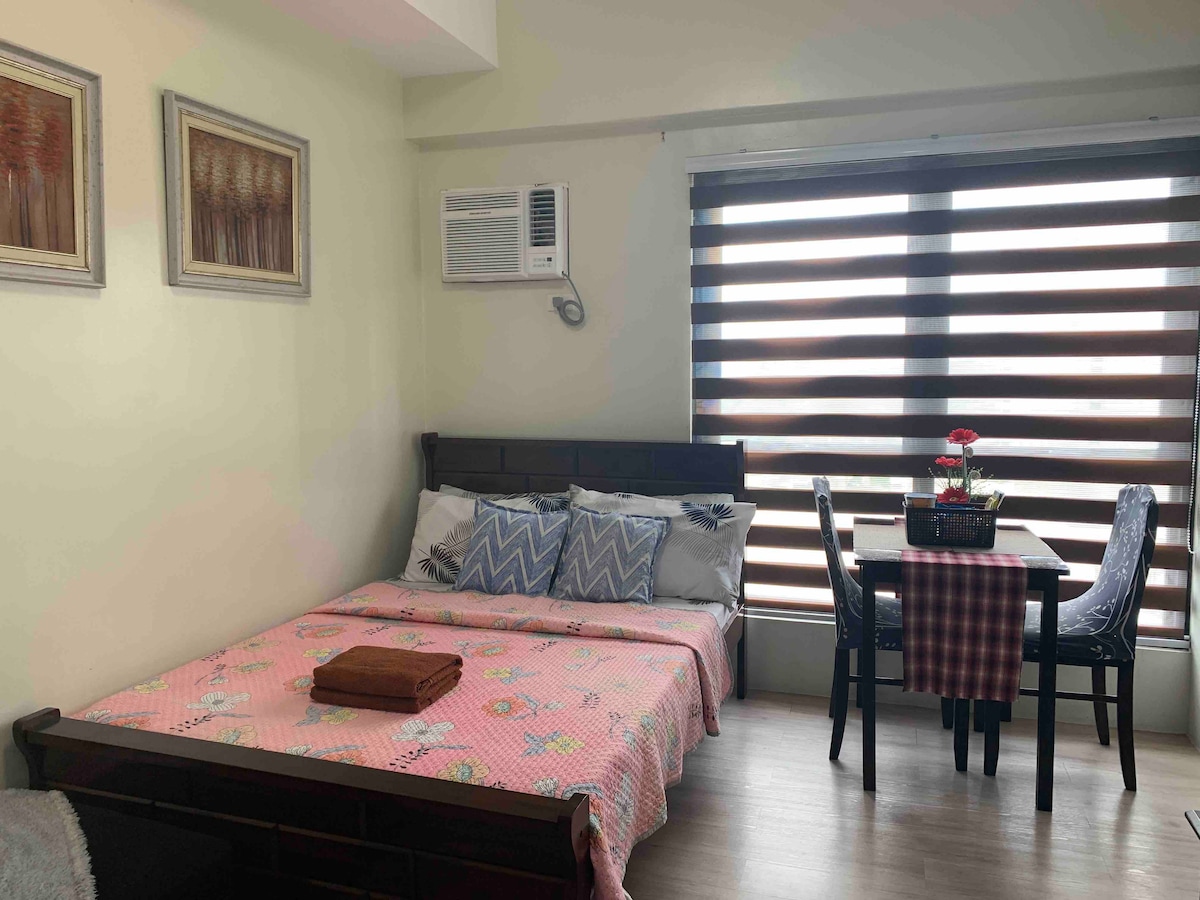 A studio space features a comfortable sleeping area with a floral patterned blanket draped over a spacious bed. Natural light enters through a window adorned with horizontal wooden slats. A small dining table is set nearby, accompanied by two chairs and a decorative centerpiece.