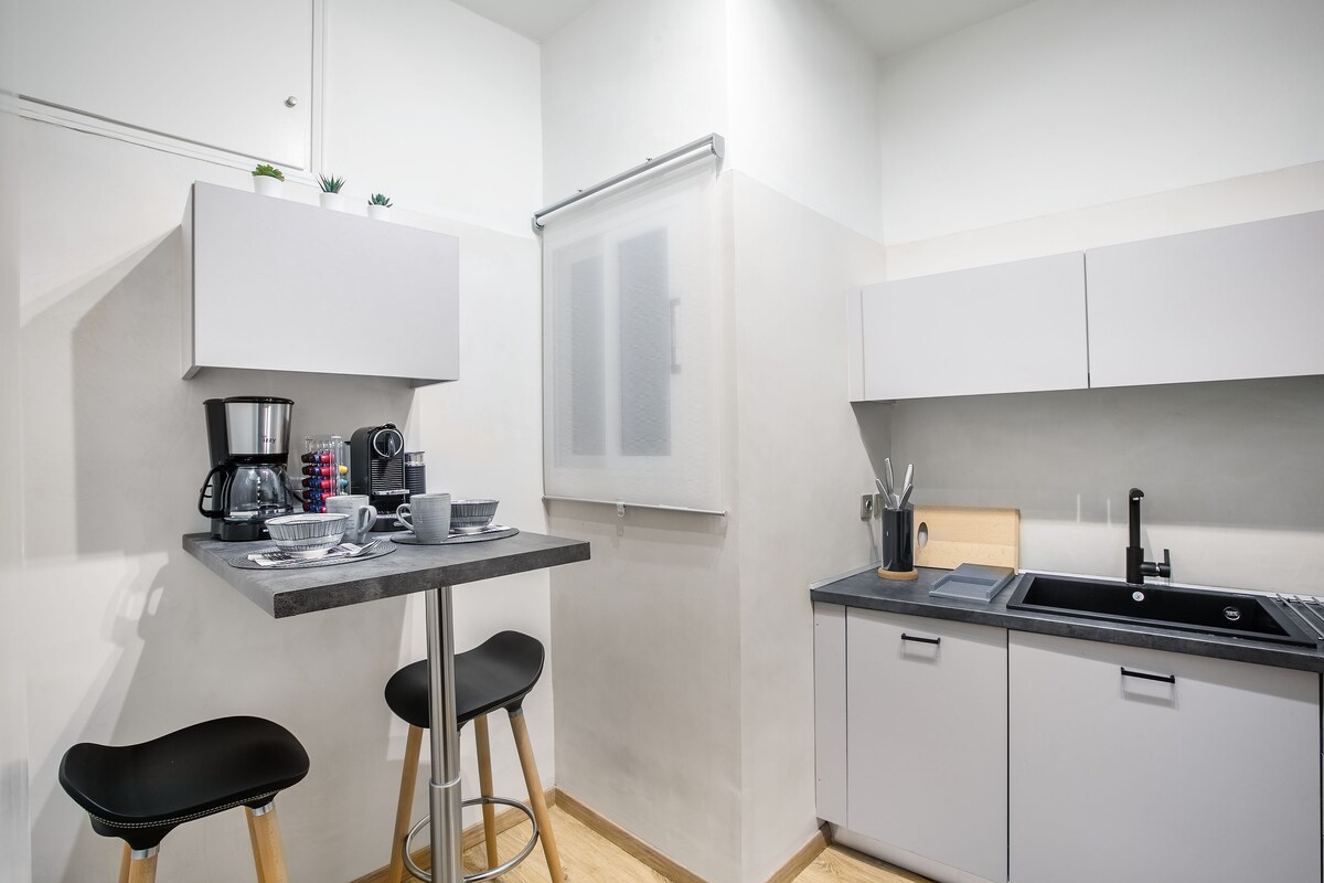 A modern kitchen is equipped with a coffee maker, a kettle, and essential cooking utensils. A compact dining table is paired with two stools, positioned near the wall. Grey cabinets and countertops provide ample storage and workspace, creating a functional cooking area.