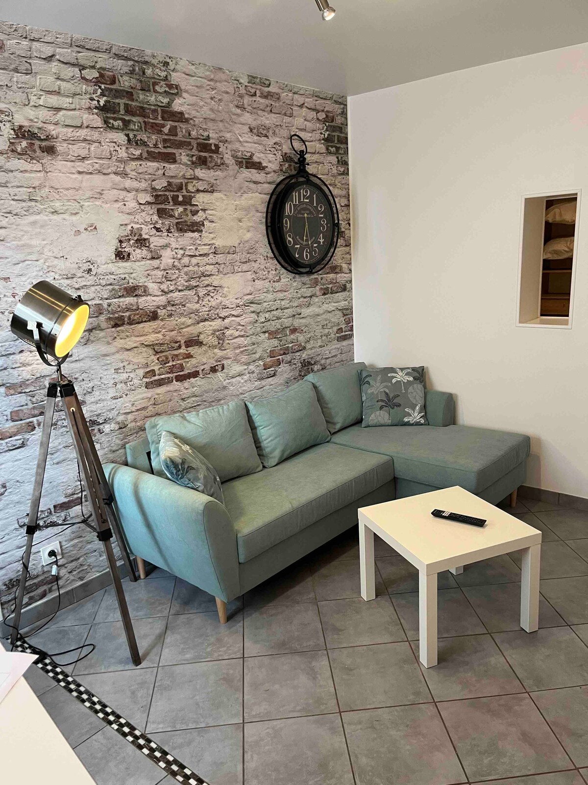 A cozy living area features a light blue sectional sofa positioned comfortably against a textured brick wall. A modern floor lamp stands beside the sofa, providing soft illumination. A small white table is placed in front, offering a space for refreshments or decor.