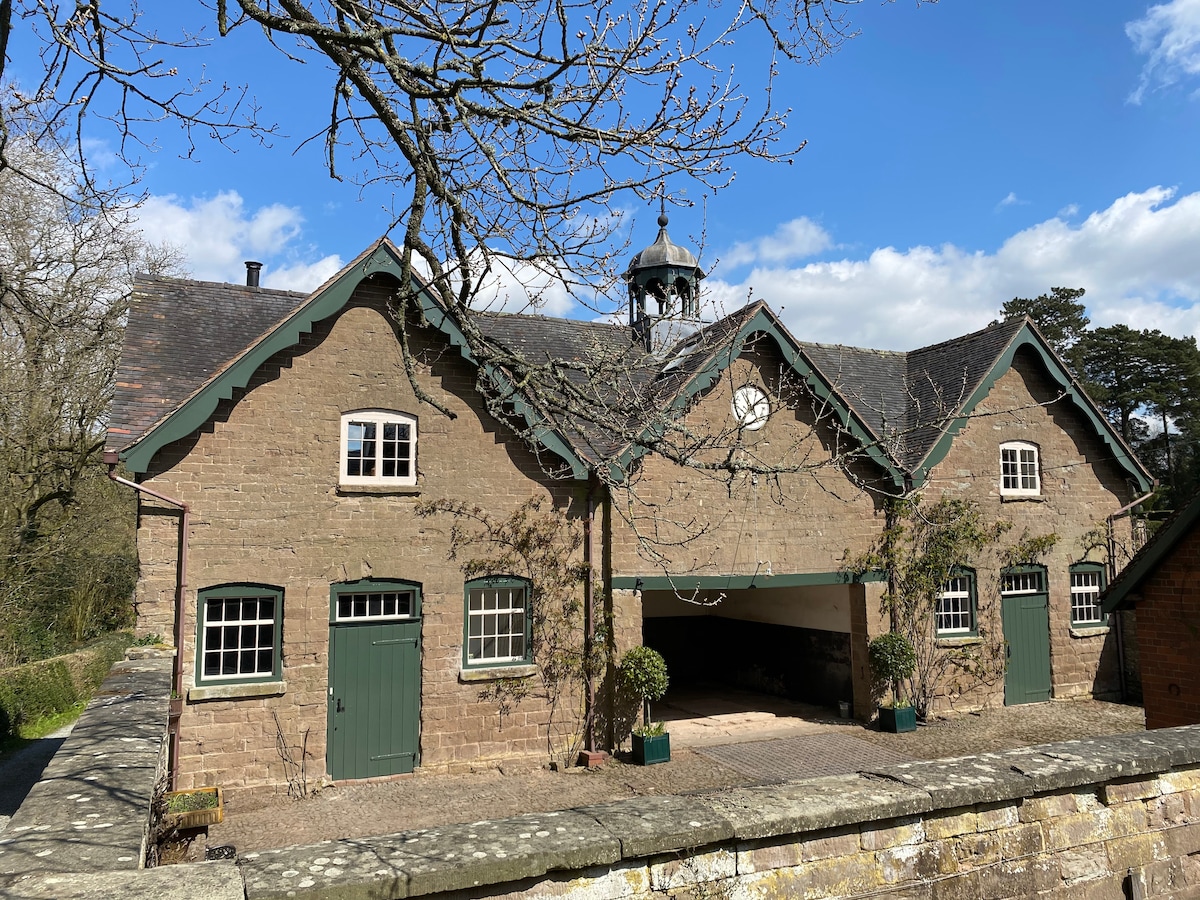 The exterior of a Victorian stable block is shown, featuring a stone façade with green accents. Windows are framed symmetrically, and a central clock tower rises above the roofline. A tree branches gently in the foreground, bordered by a stone wall enclosing the property.