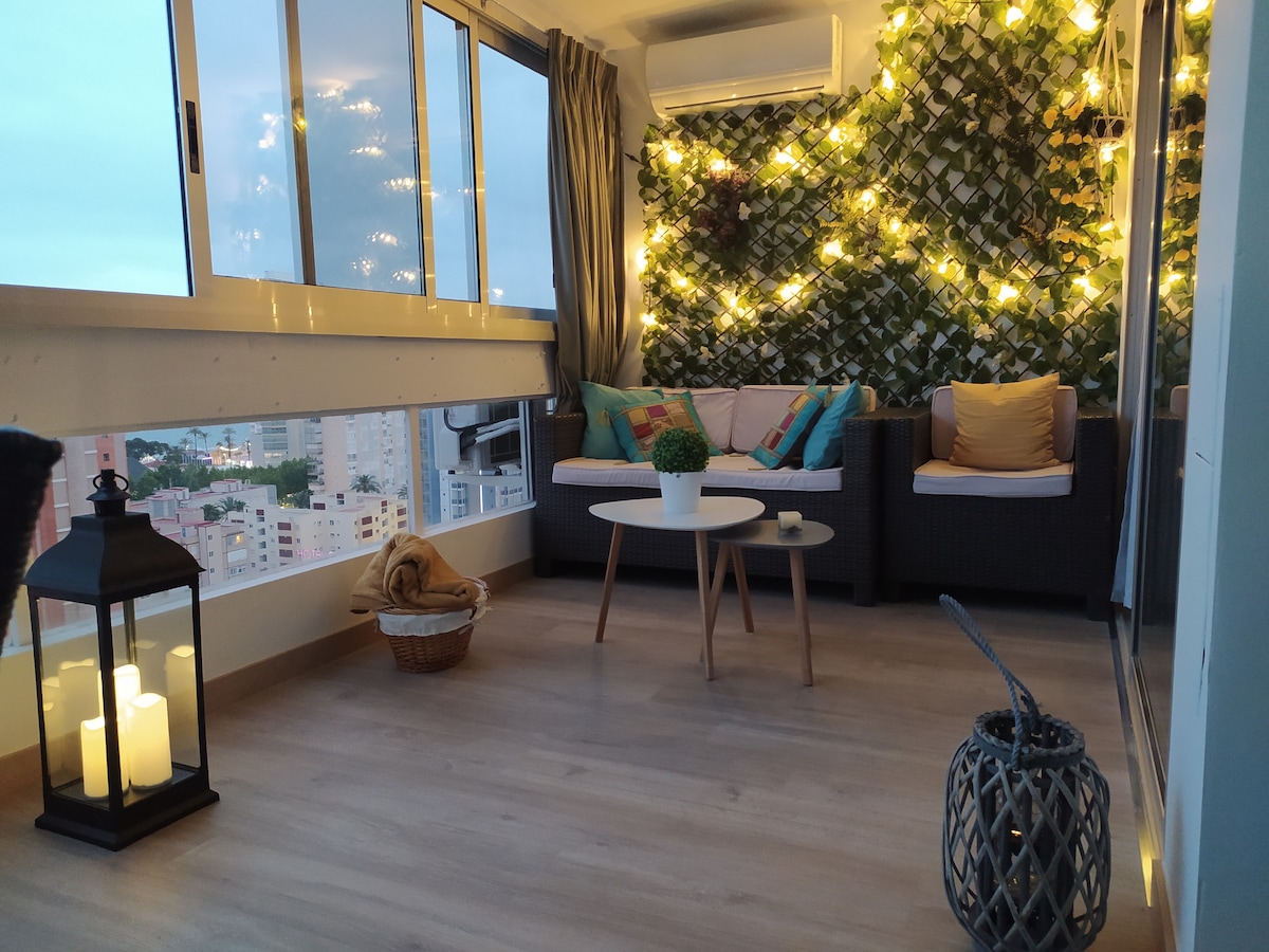 A sunlit terrace is adorned with soft lighting, creating a cozy atmosphere. Comfortable seating is arranged around a circular table. Greenery decorates the wall, and decorative lanterns and a woven basket are positioned on the floor, adding to the inviting space.
