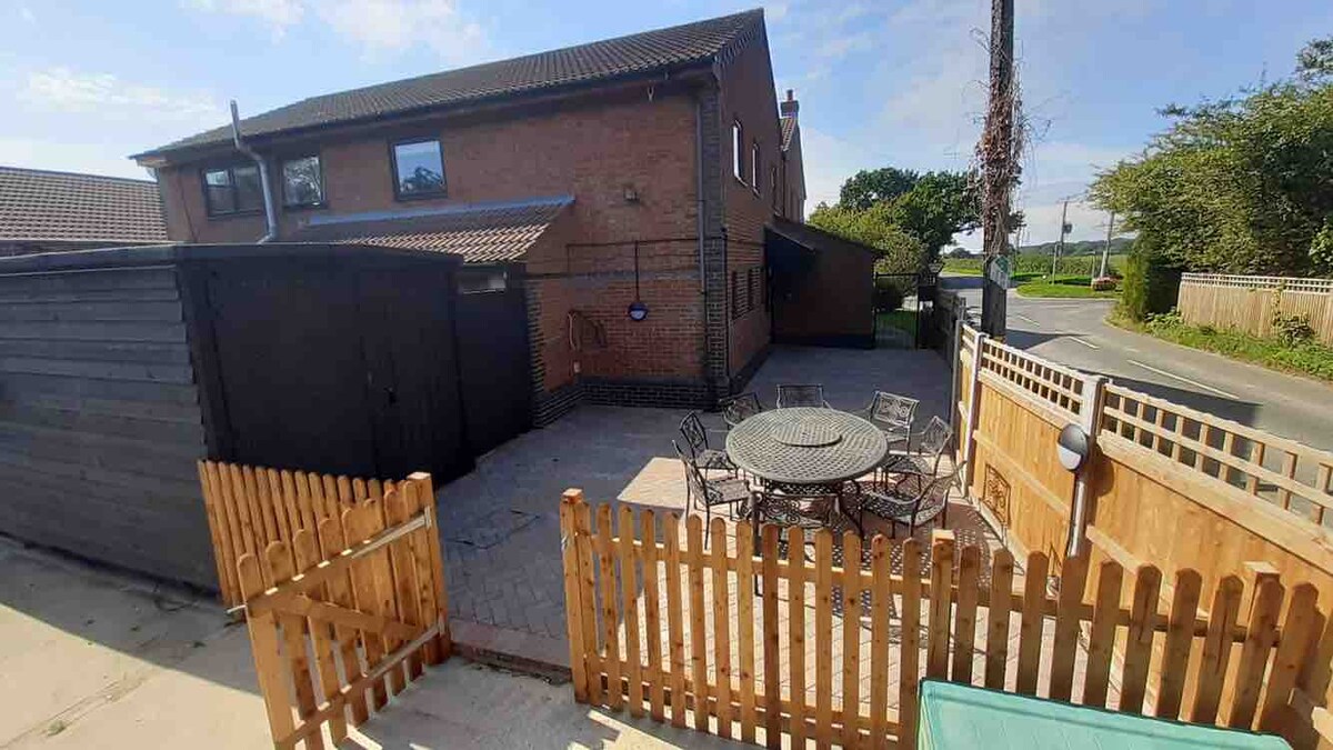 A spacious outdoor patio area is enclosed by a wooden fence, featuring a round dining table surrounded by several chairs. The area is paved and positioned against a backdrop of greenery and the exterior of the apartment.