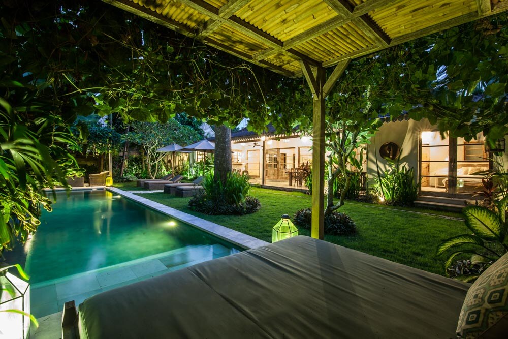 An inviting outdoor area is showcased, featuring a serene swimming pool surrounded by lush greenery. Soft lighting from decorative lanterns creates a gentle ambiance. The villa's open living space can be seen in the background through large glass doors, seamlessly connecting with the tranquil environment.