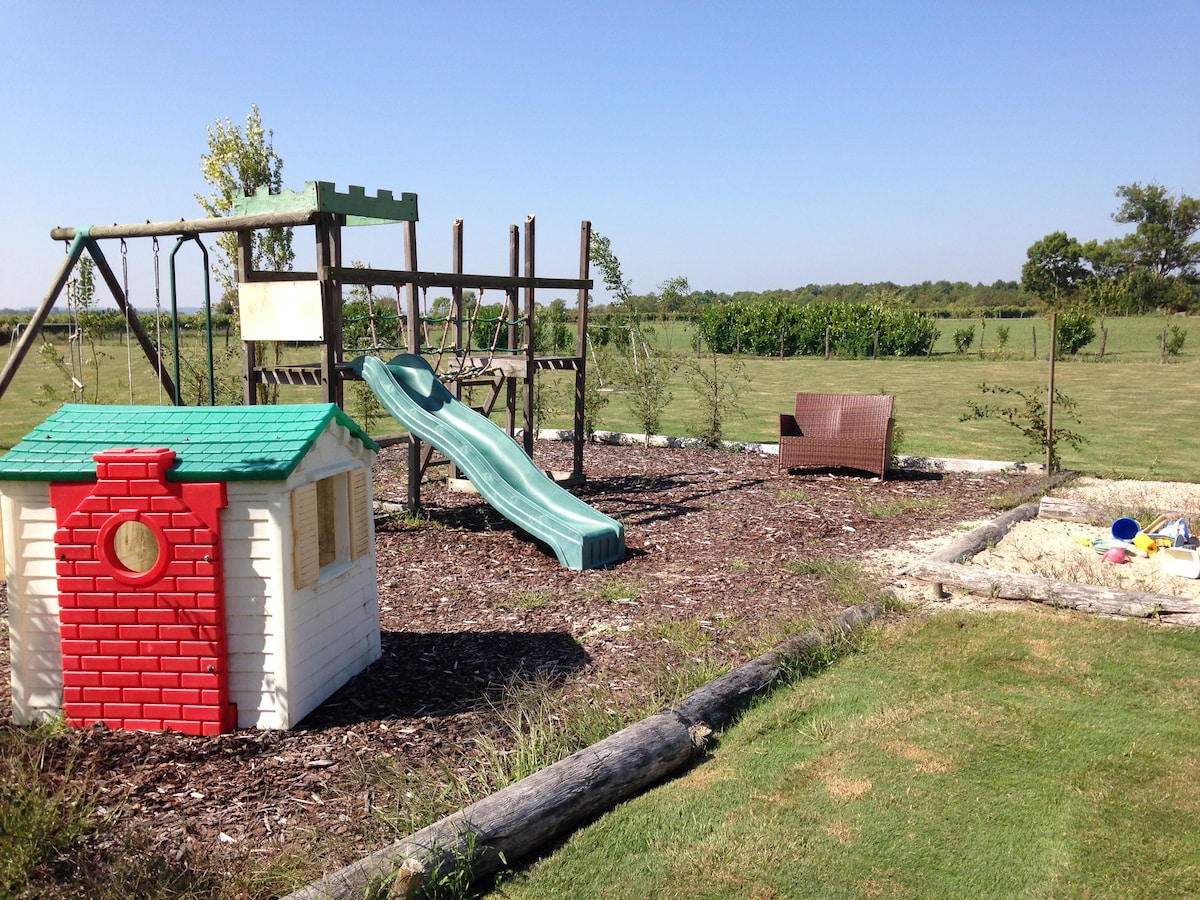 A children's play area features a colorful playhouse beside a slide and swings. A sandy play pit is visible, along with a seating area that includes a wicker chair. The backdrop showcases a spacious lawn surrounded by green vineyards under clear blue skies.