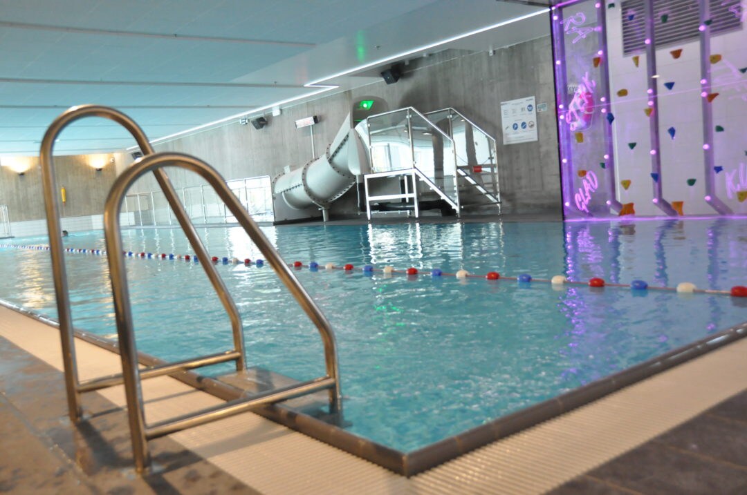 An indoor aquatic facility features a well-maintained swimming pool with a smooth entry ladder. A colorful water slide is visible alongside the pool, complemented by safety ropes marking the swimming area. Soft lighting enhances the modern design of the surroundings.