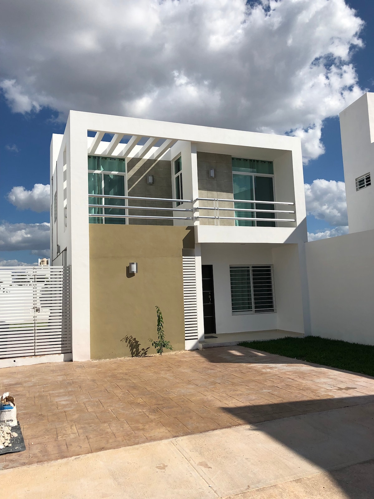 A modern two-story house features a light-colored facade with large windows and a balcony on the second floor. The exterior is complemented by a well-maintained lawn and a paved driveway leading to the entrance, under a clear blue sky dotted with clouds.