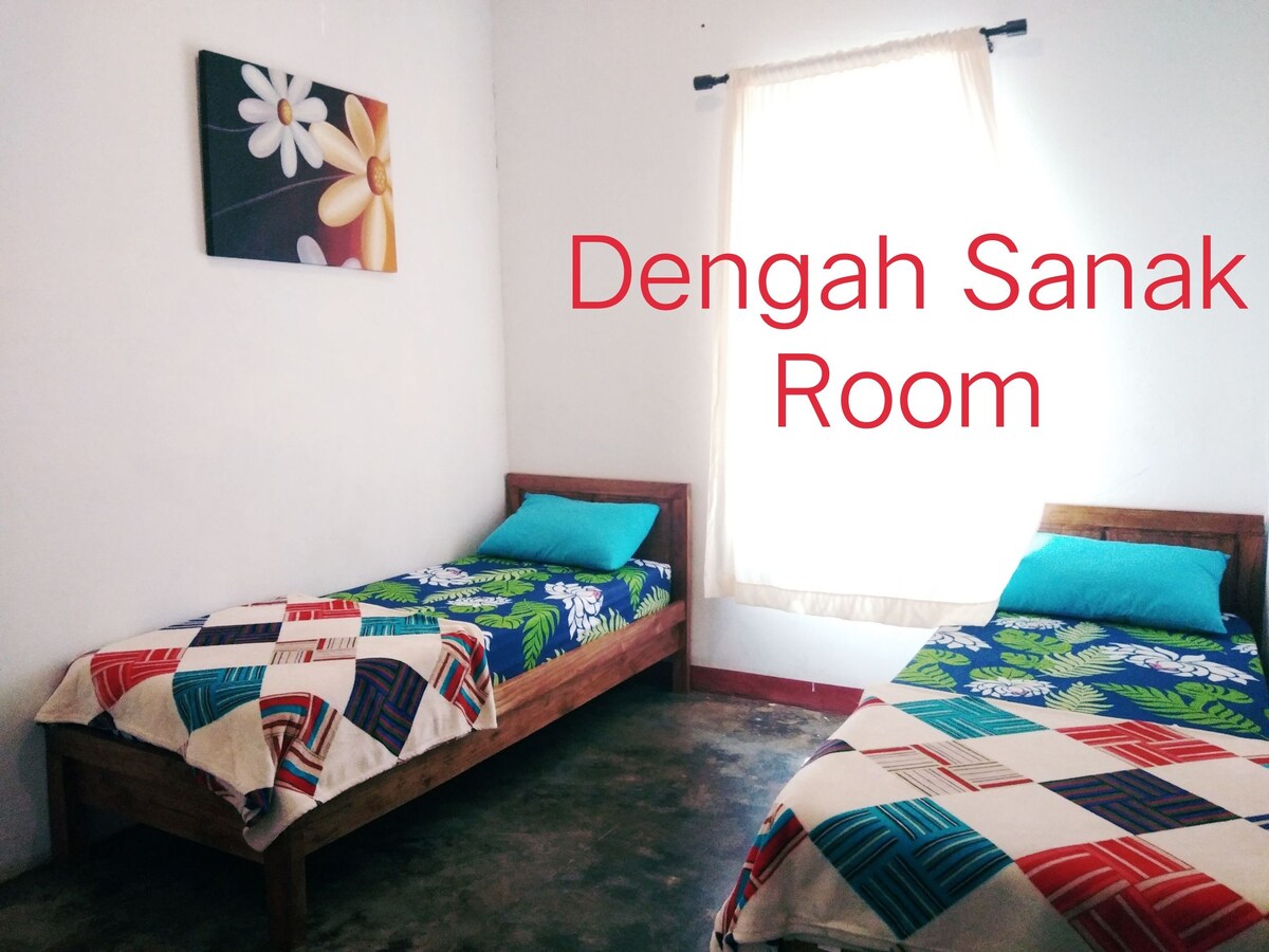 A well-lit room is equipped with two wooden beds, each adorned with vibrant floral-patterned bedcovers. Natural light enters through a large window, complementing the simple yet welcoming decor, including a framed floral artwork on the wall.