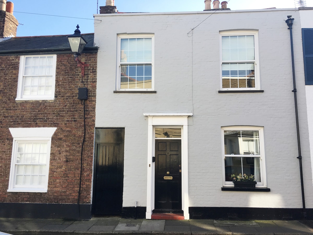A charming Georgian house appears on a quiet street. The white exterior features two large windows with multiple panes flanking a central black front door. A dark brick building is adjacent, complementing the inviting facade.