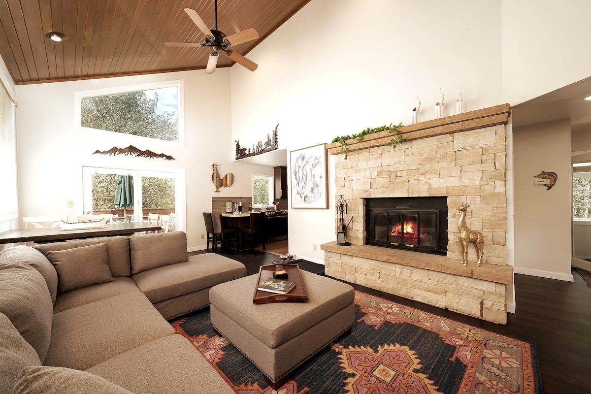 A bright living area features a large sectional couch accompanied by a square ottoman. A stone fireplace is positioned against the wall, providing a warm focal point. Dining space with a wooden table and chairs is visible through an open layout, enhancing the inviting atmosphere.