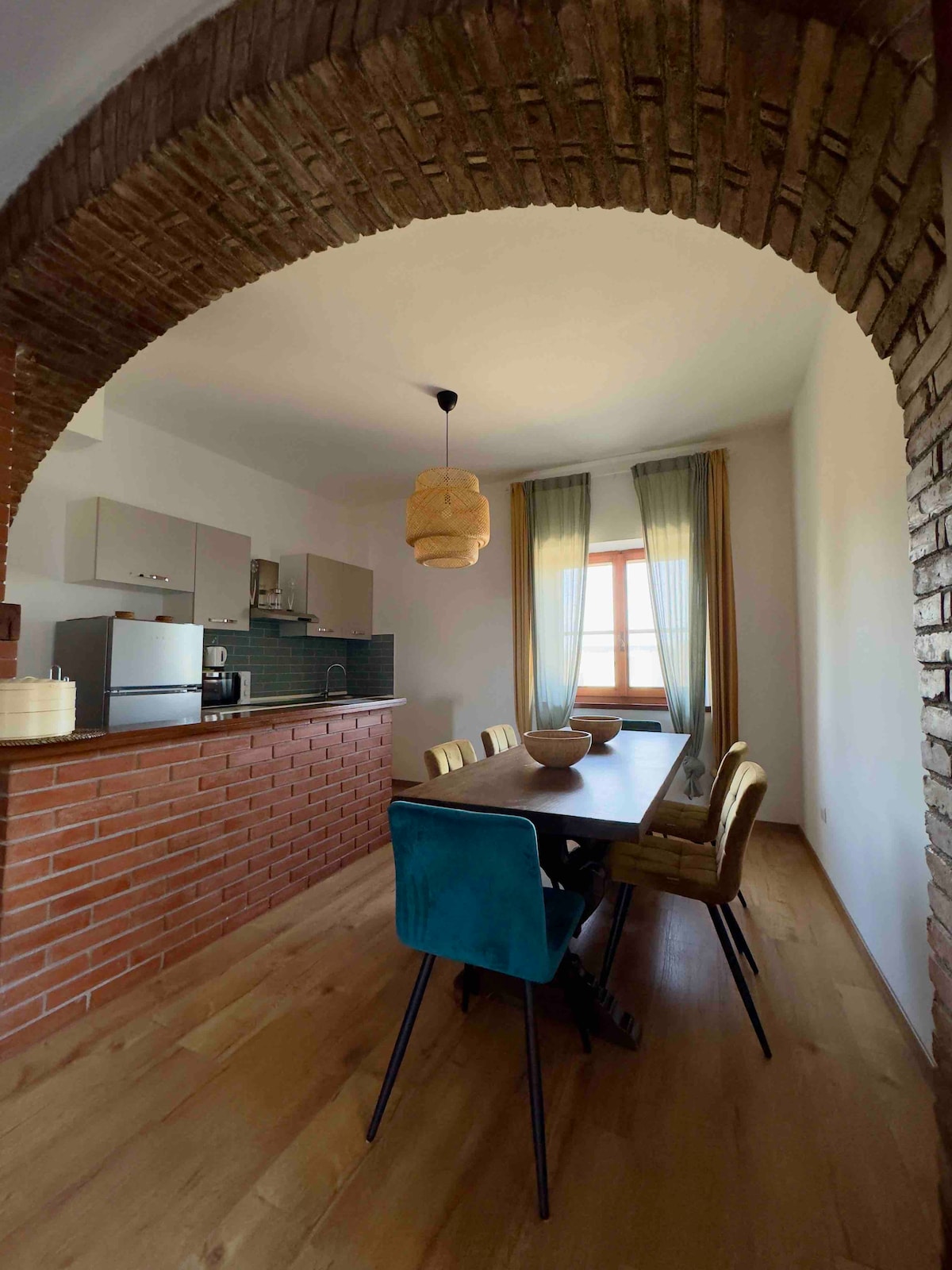 An inviting dining area is defined by a curved brick archway. A wooden table is surrounded by colorful chairs, complemented by a textured pendant light. Natural light filters in through sheer curtains, illuminating the minimalist kitchen in the background.