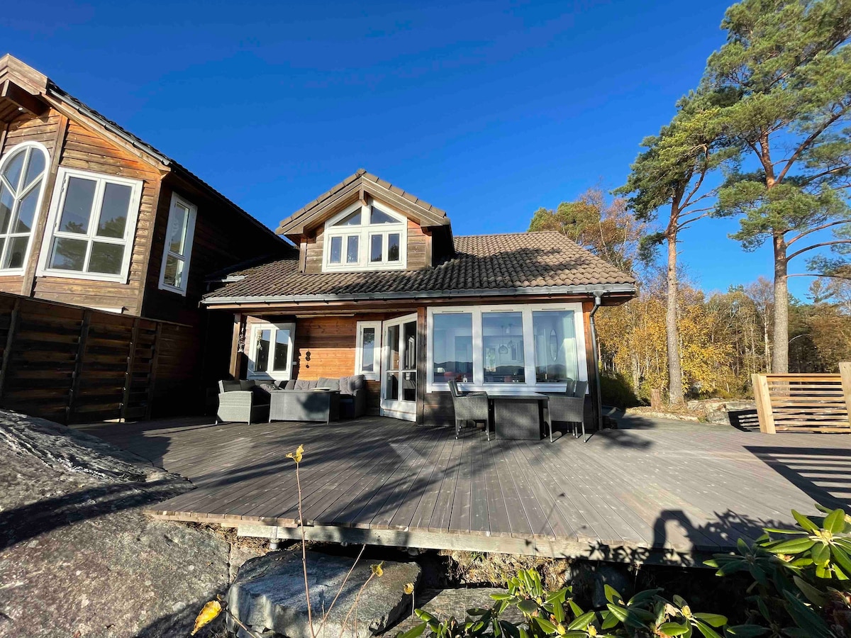 A wooden house features large windows that allow natural light to fill the living spaces. A spacious deck extends outward, furnished with outdoor seating arrangements. Surrounding trees add a touch of greenery, and the clear blue sky provides a bright backdrop.