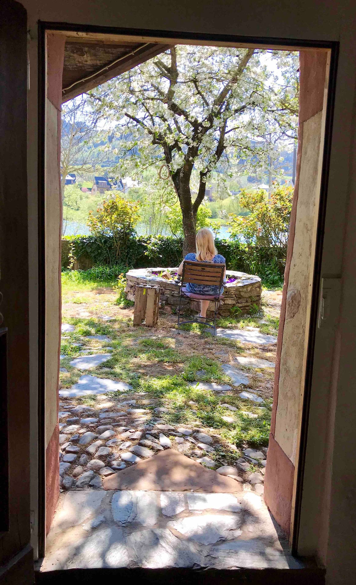 A view from an open door reveals a tranquil garden scene. A stone table is positioned beneath a flowering tree, surrounded by greenery. A person is seated with their back to the camera, enjoying the peaceful surroundings and natural light illuminating the space.