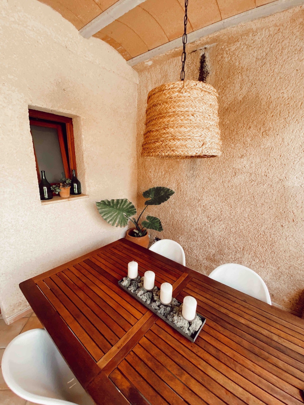 A wooden dining table is set with four white chairs, creating an inviting dining space. A centerpiece of four candles is positioned on a stone tray. Natural textures are highlighted by a woven pendant light and a potted plant, complementing the stone wall and window.