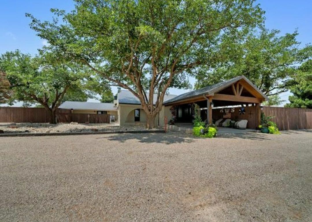 A charming exterior view of the estate is framed by large, mature trees. The entrance features a covered porch with wooden beams and a gravel driveway leading to the front door, creating an inviting first impression.