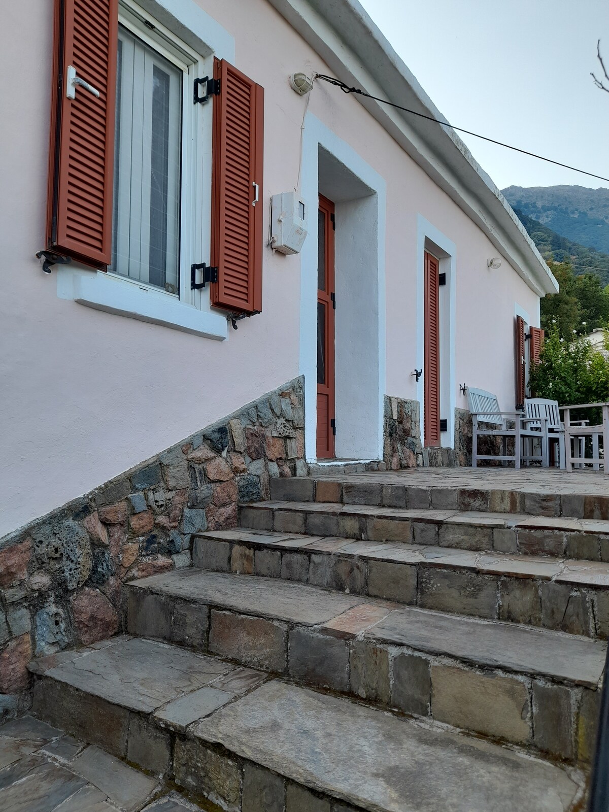 A stone pathway leads up to a charming pink house with red shutters and a wooden door. The entrance is framed by a textured stone wall and is flanked by outdoor seating. Natural light highlights the steps and surrounding greenery.
