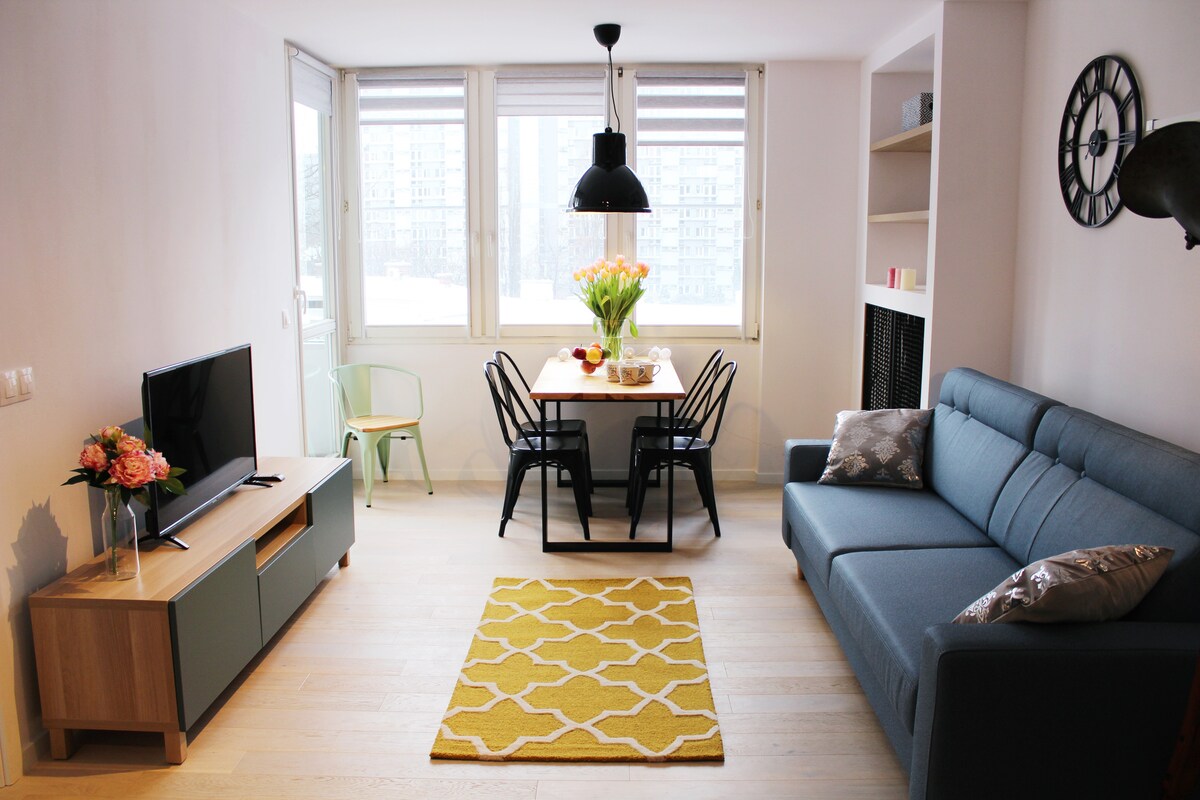 A bright and open living area features a comfortable blue sofa and a wooden entertainment unit with a television. A dining table with black chairs is set for meals, accented by a vase of tulips. Large windows provide natural light and a view of the city.