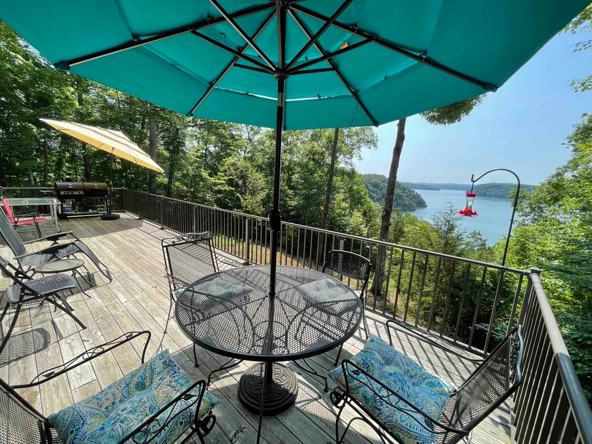 An outdoor deck is furnished with various seating options, including a circular glass table and several black metal chairs. An umbrella provides shade, while a grill is positioned in the background. The deck offers an expansive view of the lake and surrounding trees.