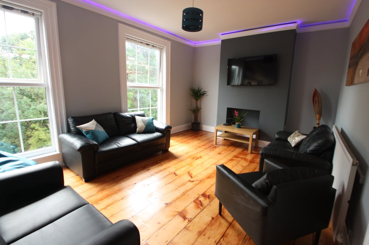 A spacious living room features two leather sofas facing each other, with cushions in shades of teal. Natural light fills the room through large windows, showcasing wooden flooring. A wall-mounted television is positioned above a modern coffee table, with a decorative plant placed nearby, enhancing the atmosphere.