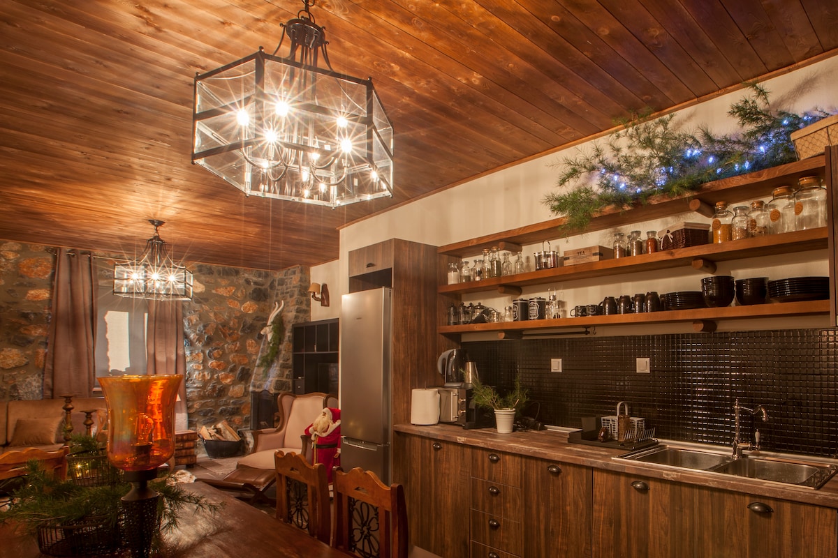 The kitchen area features natural wood cabinetry and shelves displaying glassware and dishes. Modern appliances, including a refrigerator and kettle, are visible. Soft lighting from stylish fixtures illuminates the space, complementing the stone accent wall and rustic wooden ceiling.