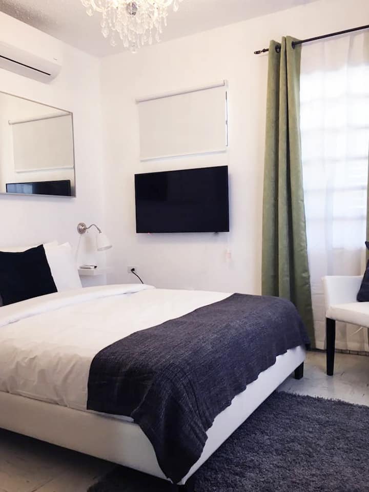 Vista del cuarto de habitación el cual incluye toallas y ropa de cama,mesa de noche con lámpara, closet, butaca, TV con sistema ROKU, Internet y una cama Queen para 2 personas. 