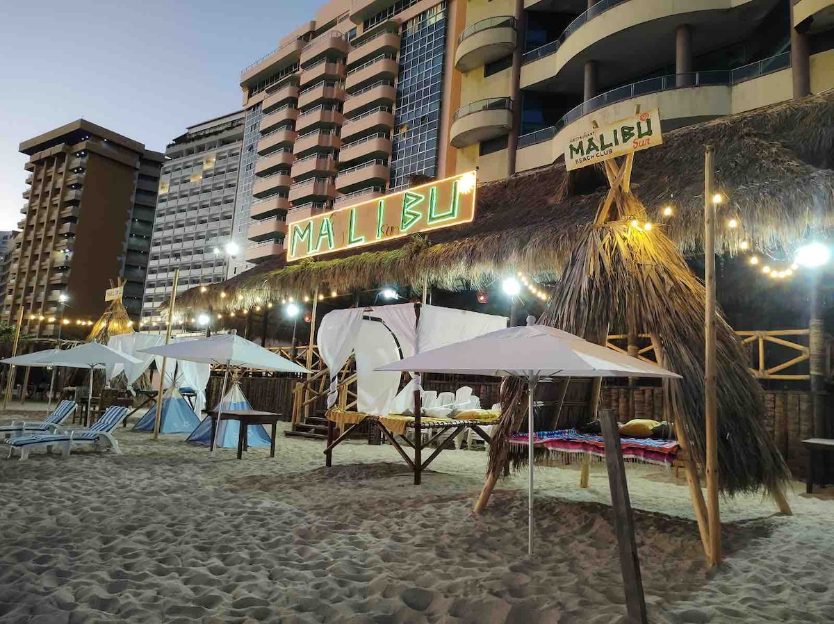 A beach area is depicted with several lounge chairs and shaded cabanas under a thatched roof. Colorful lights outline a sign that reads 'MALIBU.' The beachfront setting features soft sand and nearby tall buildings, creating a vibrant atmosphere for relaxation.