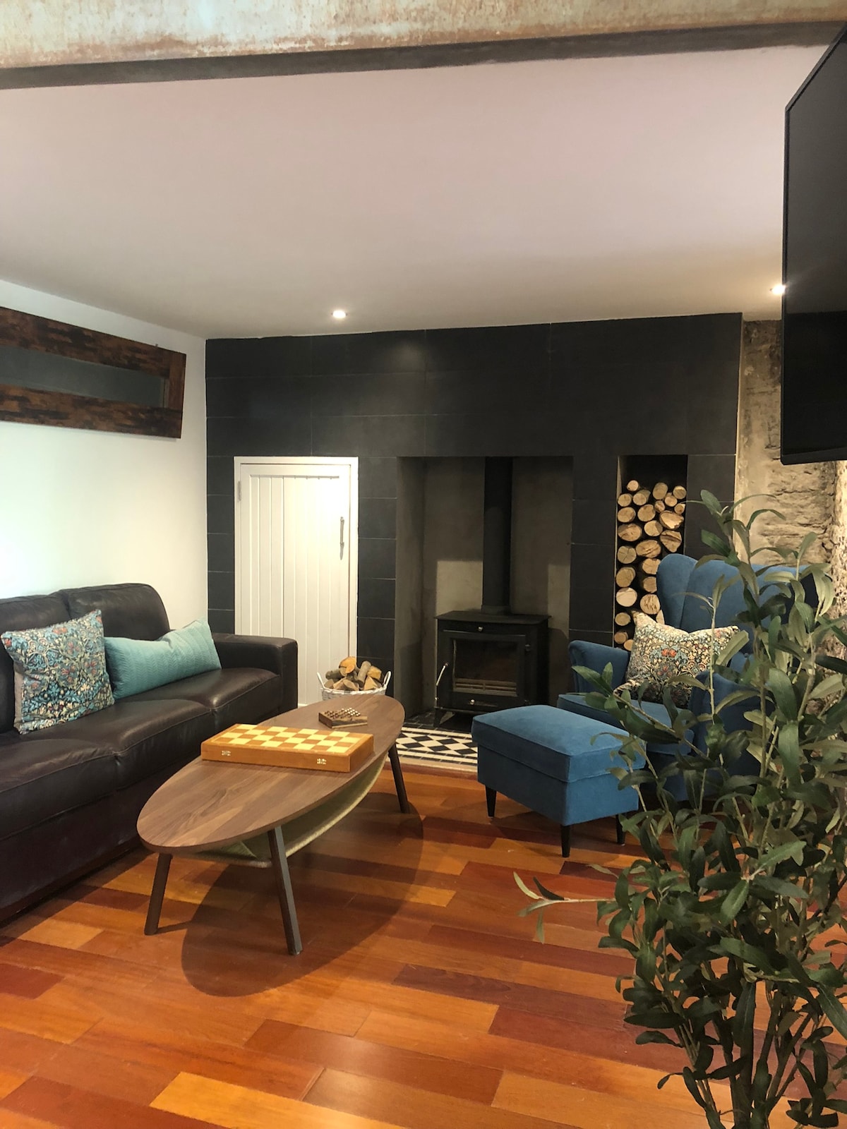 A cozy living room features a dark leather sofa accompanied by a blue armchair. A stylish coffee table is positioned in front, and a wood-burning stove is set against a black tiled wall. A stack of logs is neatly arranged nearby.