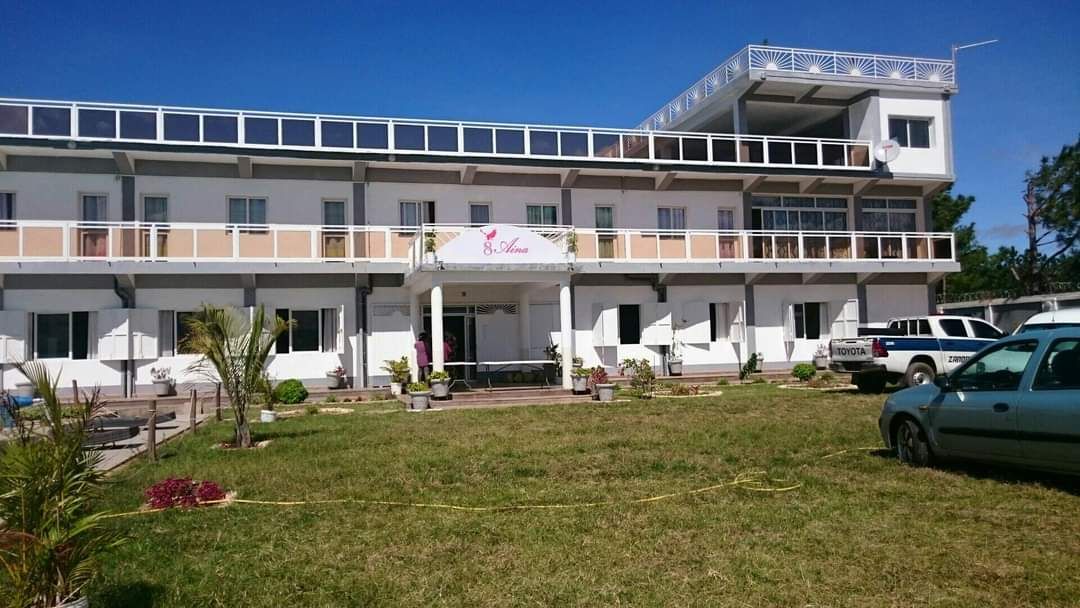 A modern, multi-story building is shown, featuring a white facade and large windows. The entrance is highlighted by a sign, while surrounding greenery includes small planted areas. Several vehicles are parked nearby on a grassy area that adds to the serene setting.