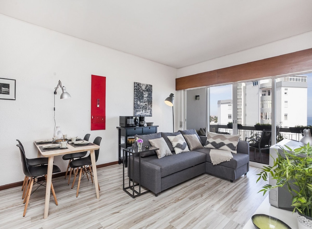 A modern living area showcases a spacious grey sectional sofa, complemented by a black coffee table and a dining table for four. Large windows allow natural light to flood the room, offering views of the surrounding buildings and balcony access.