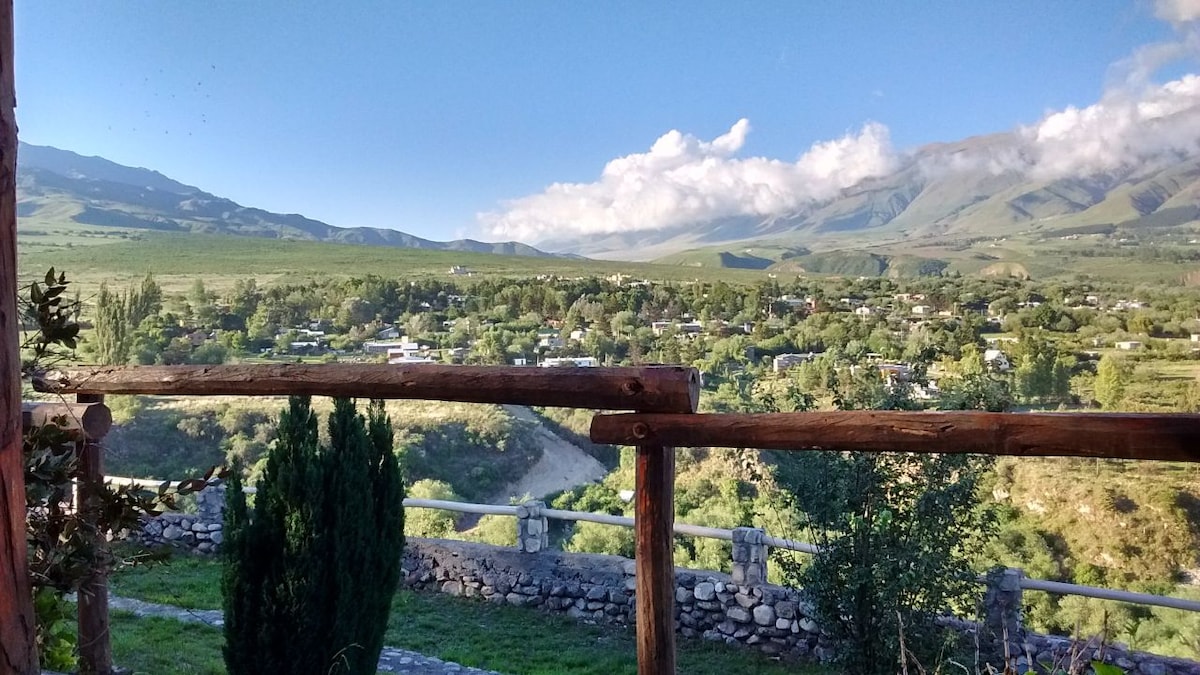 A panoramic view of a lush green valley framed by mountains under a clear blue sky. A wooden railing overlooks the landscape, highlighting the trees and scattered homes in the distance. Natural light enhances the scenery, inviting relaxation and appreciation of the surroundings.