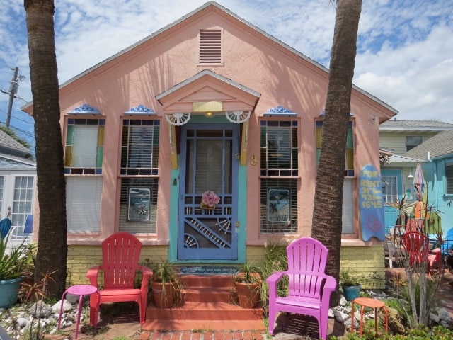 The cottage exterior features a charming pink facade with decorative accents and a welcoming front entrance. Two vibrant pink chairs are positioned on the brick pathway, framed by lush greenery. Large windows provide ample natural light, while colorful artistic details add character.