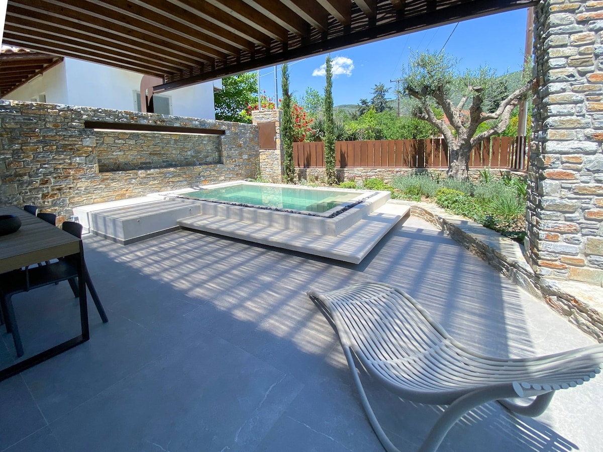 A shaded outdoor area features a small jacuzzi surrounded by stone walls and lush greenery. A sleek lounge chair is positioned nearby on a tiled floor, offering a space for relaxation. Sunlight filters through the overhead beams, creating a patterned effect on the ground.