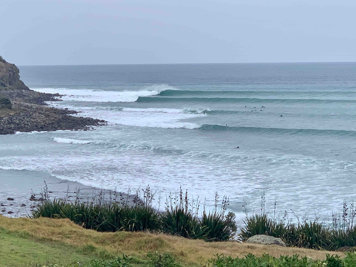 A serene coastal view features gentle waves breaking on a sandy shoreline. The ocean is calm with rhythmic swells, while surfers are seen enjoying the water. Lush green grass and coastal plants frame the foreground, complementing the tranquil scene.