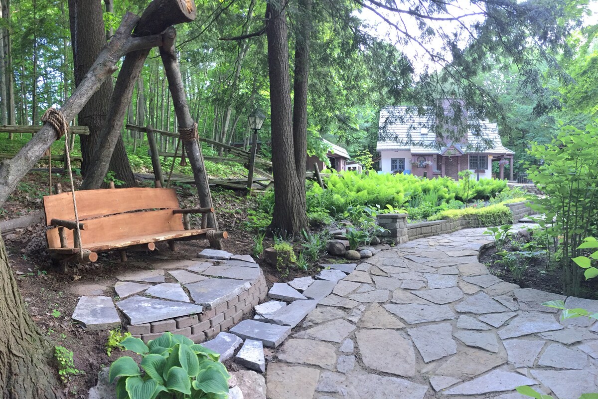 A stone pathway winds through a lush green garden, bordered by ferns and trees. A wooden swing hangs gracefully from a sturdy tree branch, inviting relaxation. In the background, a charming cottage is partially obscured by foliage, creating a serene and natural setting.