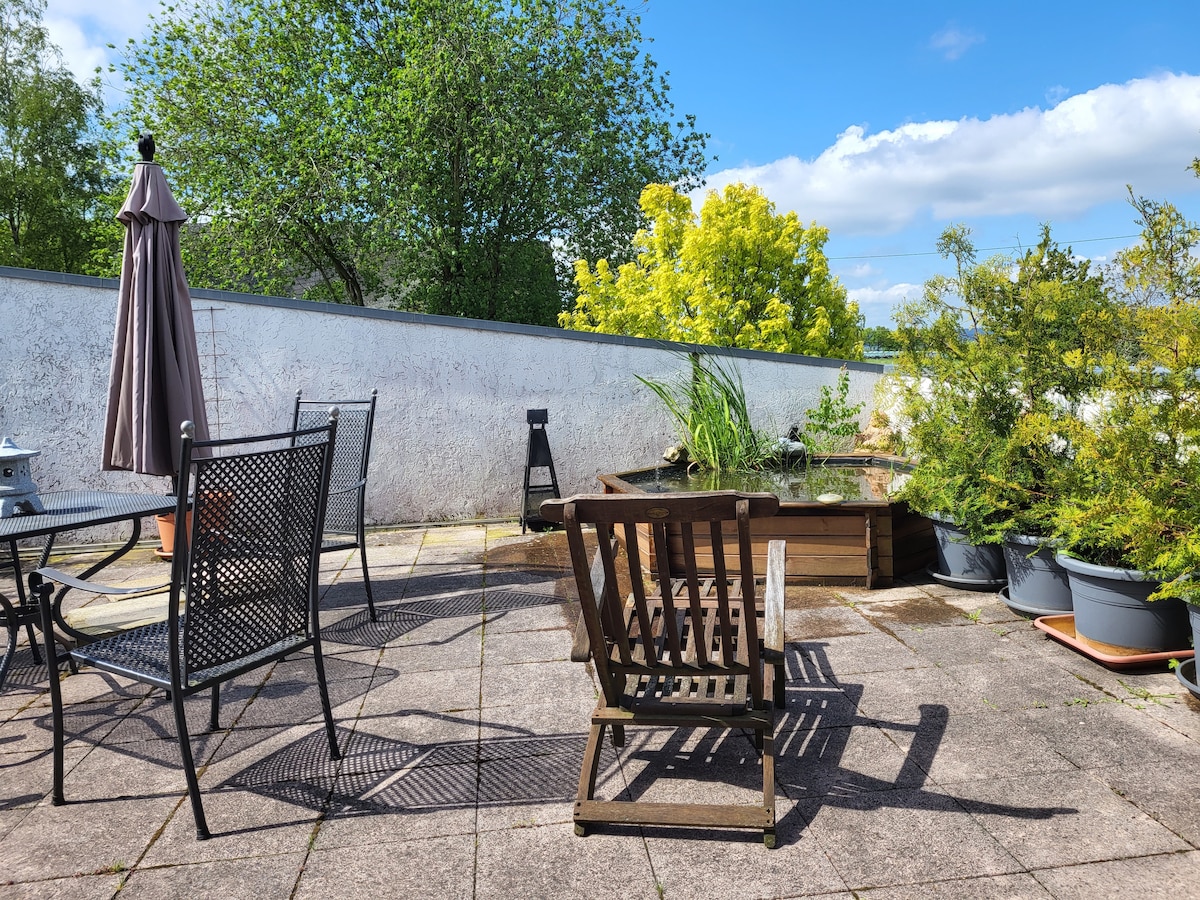 An outdoor patio area is featured with a shaded seating arrangement, including a wooden chair and metal tables. Potted plants and a small wooden garden bed add greenery, while a wall reflects bright sunlight and a clear blue sky.