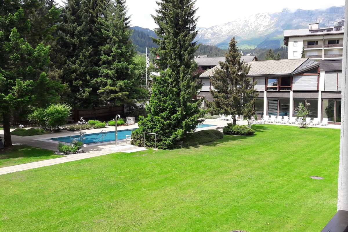 The image depicts a well-maintained grassy area surrounded by trees, with two heated pools visible at the center. Sun loungers are arranged nearby, and a scenic mountain backdrop can be seen in the distance under a clear sky.