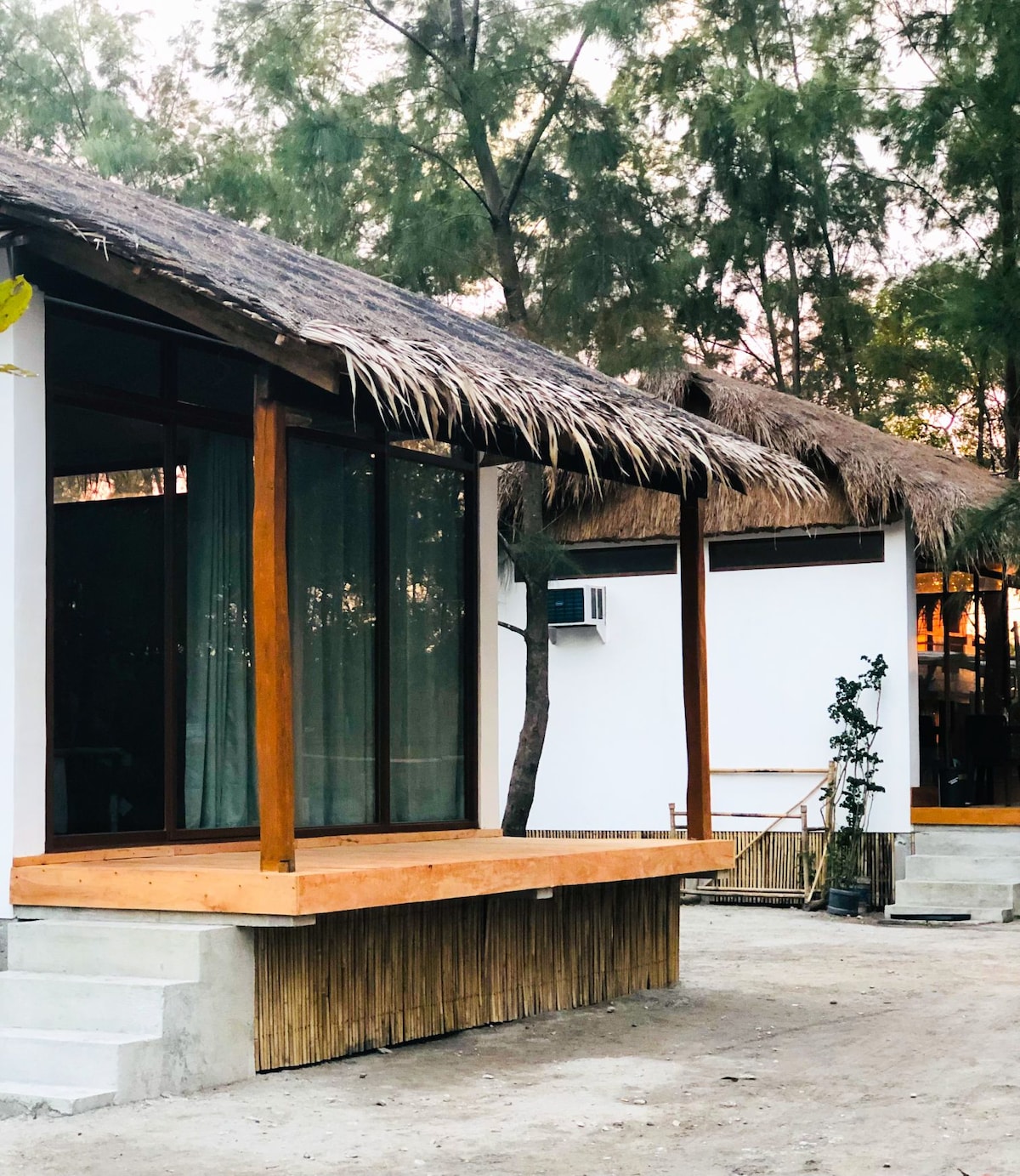 A series of charming, thatched-roof structures are set among trees, with wooden decks extending out from the front. Large glass windows allow natural light to fill the space, while a small air conditioning unit is visible on the side of one unit.