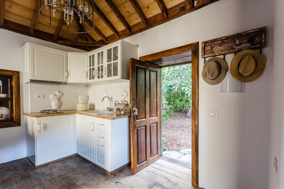 A compact kitchen area features white cabinetry and a wooden countertop. Natural light is welcomed through the open door, leading to a garden. Hat racks are positioned near the entrance, and decorative items are displayed in a nearby cabinet.