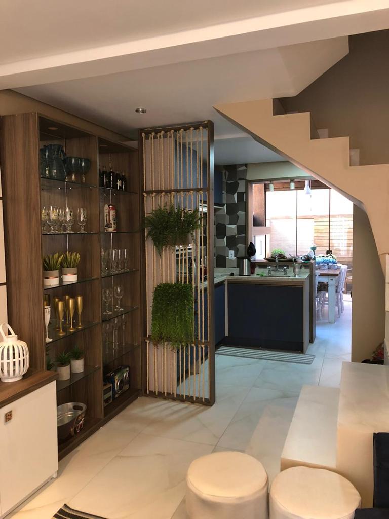 An open living area showcases a modern design with a wooden shelving unit displaying glassware and decorative plants. A staircase leads to an upper level, while a glimpse of the kitchen and dining area can be seen in the background, featuring contemporary furnishings.
