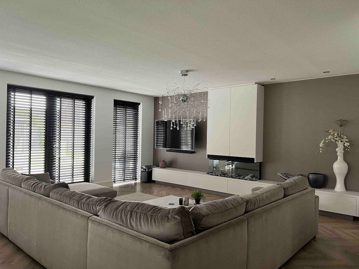 A spacious living area is presented with a large, L-shaped sectional sofa and a modern glass coffee table. A contemporary chandelier hangs from the ceiling above. Floor-to-ceiling windows with black shutters allow natural light to fill the space, complemented by a sleek media unit and decorative accents.