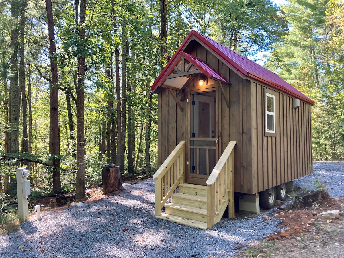 The exterior of the tiny home showcases a wooden facade with a striking red roof, complemented by a small porch with wooden steps. Surrounded by trees, the gravel path leads to the entrance, providing a natural and rustic setting.