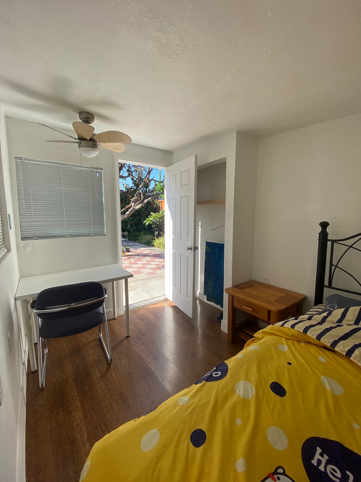 A clean and comfortable room features a spacious bed with yellow bedding and a small wooden nightstand beside it. A desk and chair are positioned near the window, which allows natural light to enter. A door leads to an outdoor space with tiled flooring.