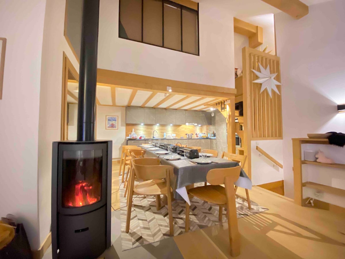 A spacious living area features a dining table surrounded by light wood chairs, complemented by a warm wood-burning stove. Soft lighting enhances the warm tones of the decor. An open staircase leads to a mezzanine level, with a star-shaped decorative element on the wall.