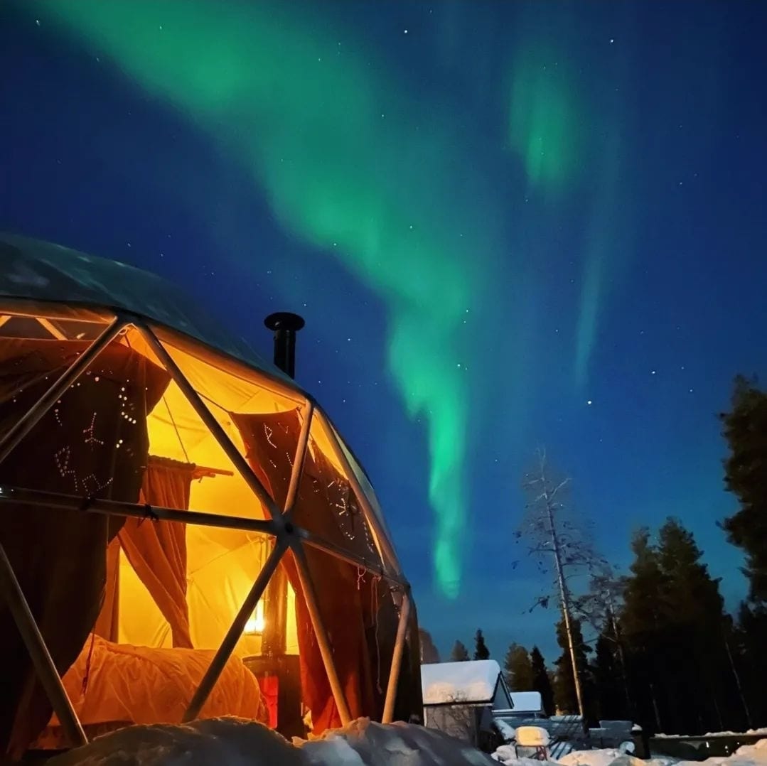 A unique igloo structure is illuminated softly from within, with the northern lights shimmering above. The clear night sky showcases vibrant green colors, while snow-covered surroundings enhance the serene atmosphere of this cozy retreat.