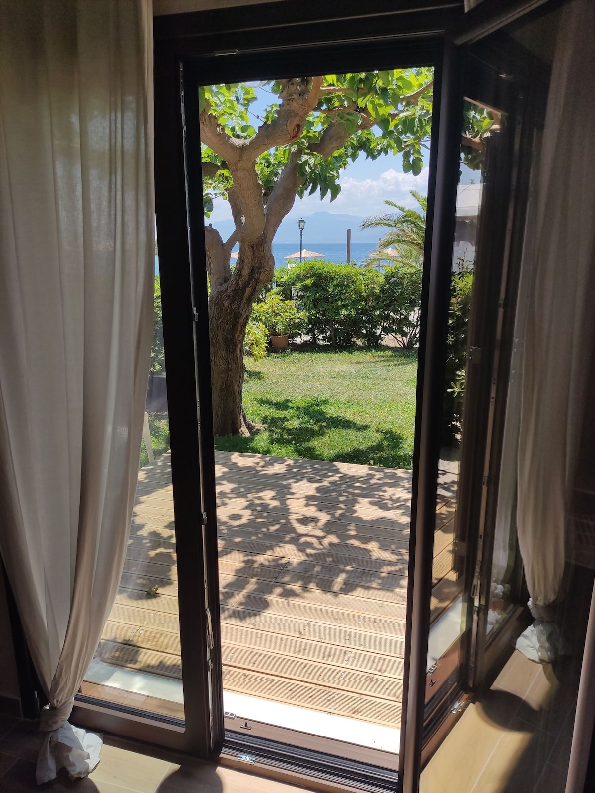 A glass door opens to a view of a wooden deck surrounded by greenery. Outside, a tree provides shade, and the sea is visible in the distance. Soft drapes frame the doorway, allowing natural light to illuminate the interior.
