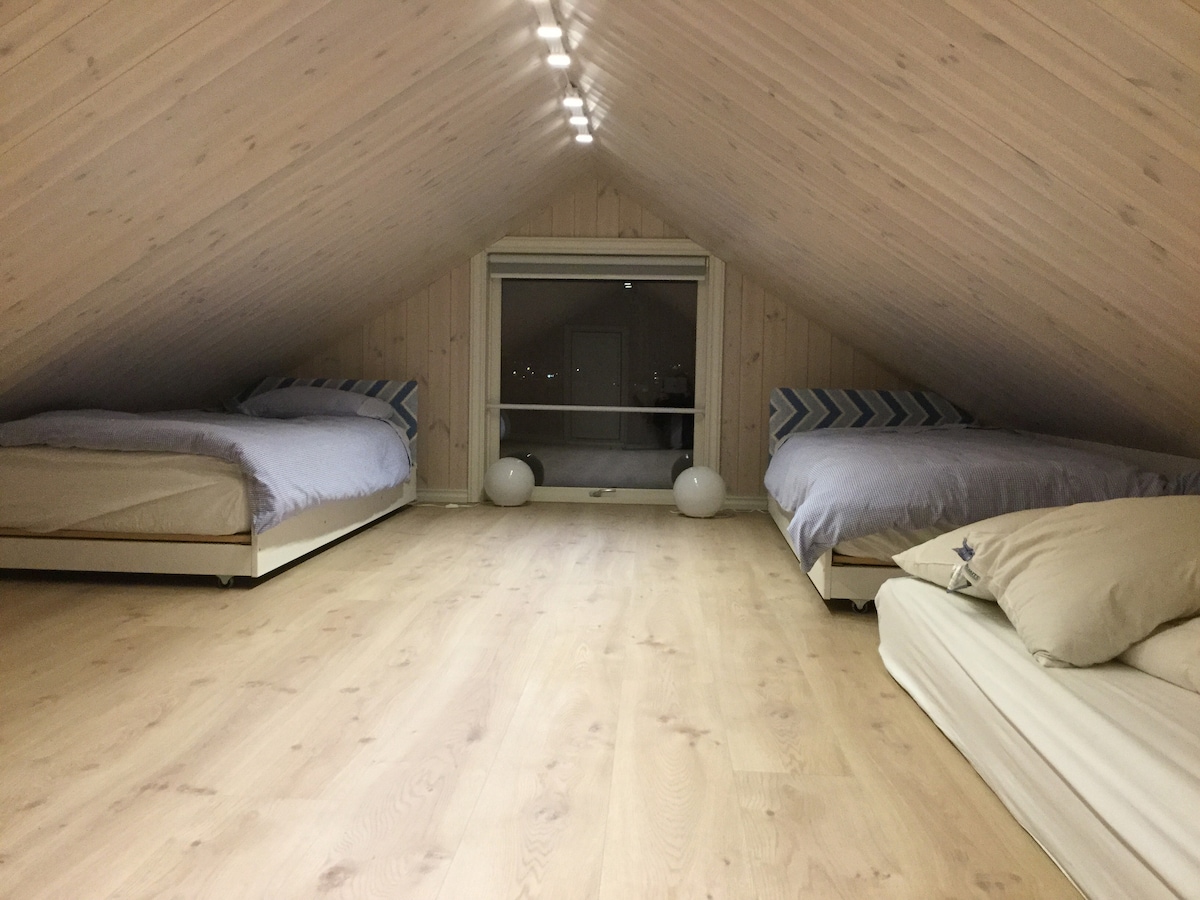 An upper loft area is presented with three spacious beds, each positioned against sloped wooden walls. A large window allows natural light to filter in, illuminating the light wood flooring. Simple decorative elements, including subtle patterned bedding, enhance the serene environment.