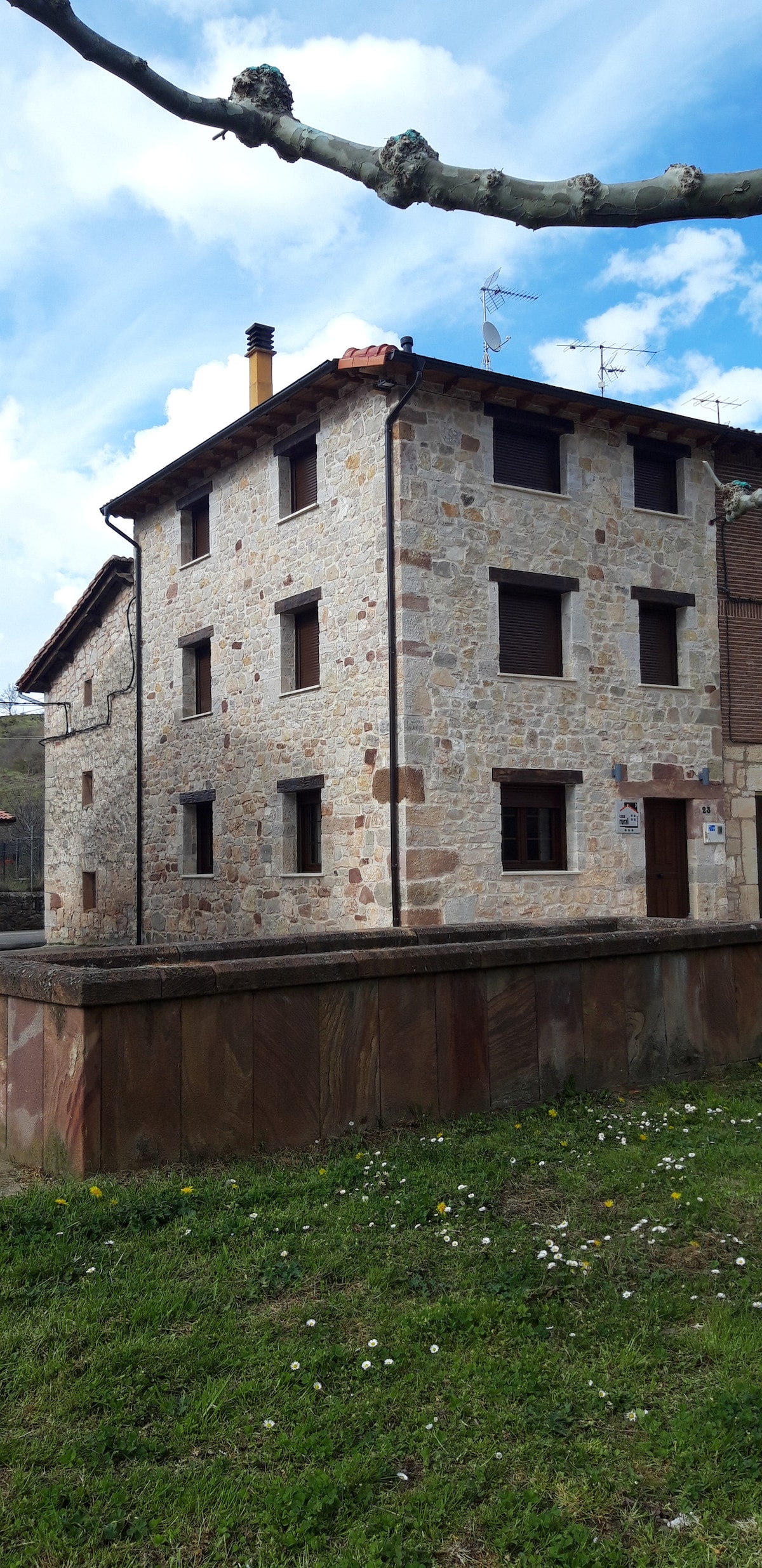 A traditional stone building is depicted, showcasing a three-story structure with multiple windows and a wooden door. The exterior is made of natural stone, and the surrounding area features a grassy lawn with wildflowers. The sky is partly cloudy, adding to the serene atmosphere.